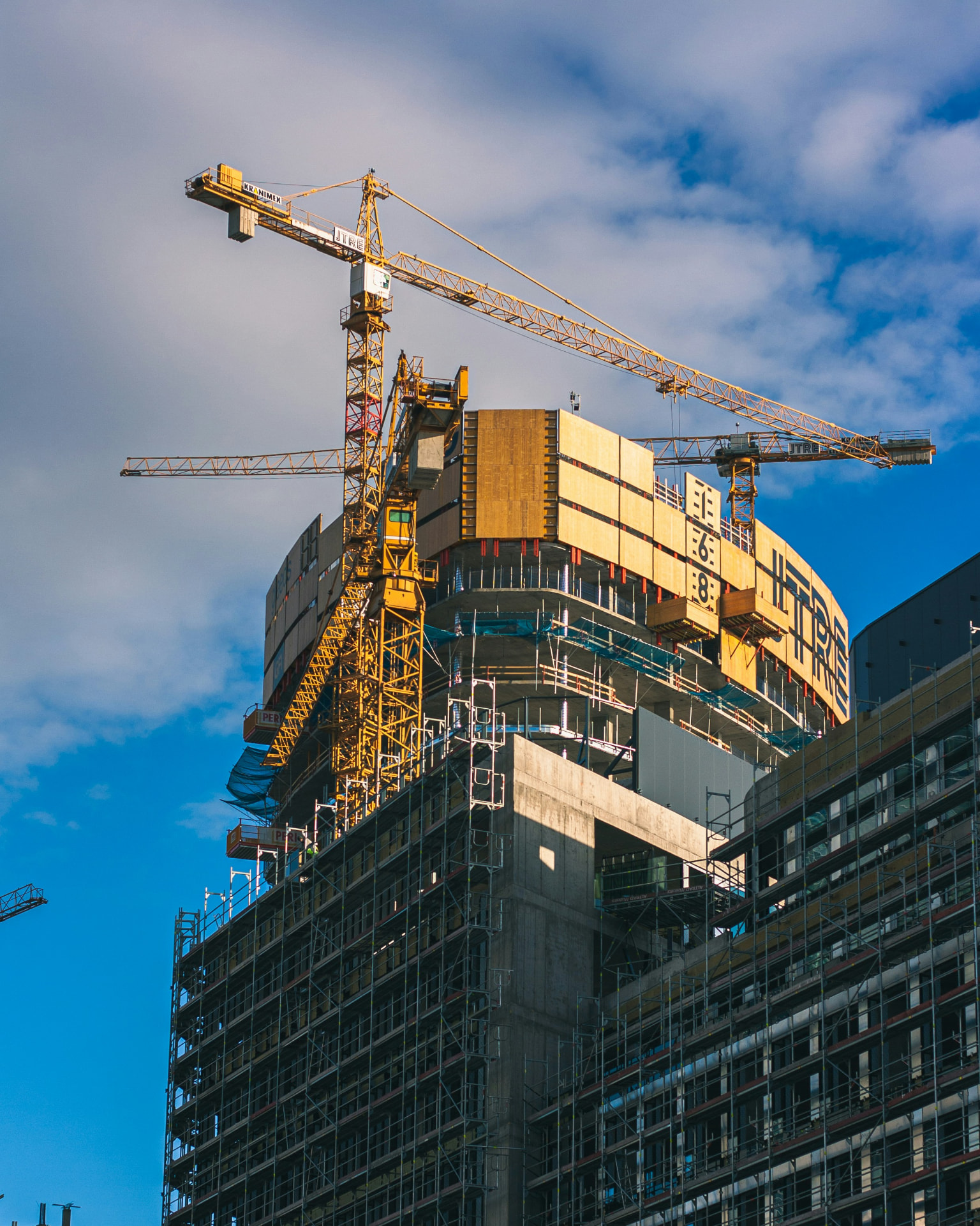 Tower cranes above a high-rise under construction, wrapped in scaffolding and climbing formwork panels.